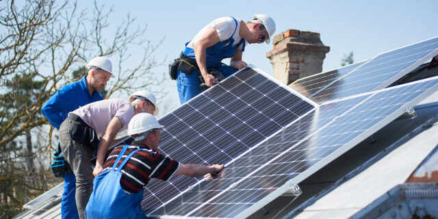 Male workers installing solar photovoltaic panel system. Group of electricians mounting blue solar module on roof of modern house. Alternative energy ecological concept.